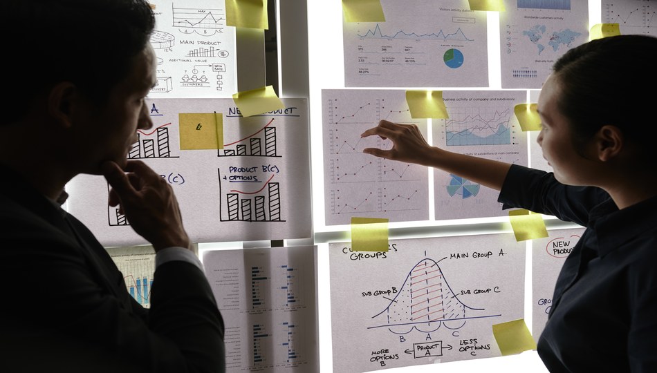 Two colleagues analyzing business charts and data visualizations on a wall, discussing strategies to overcome application modernization challenges