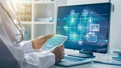 Doctor holding a tablet while viewing a healthcare data network with patient icons and analytics on a desktop monitor, representing healthcare software modernization