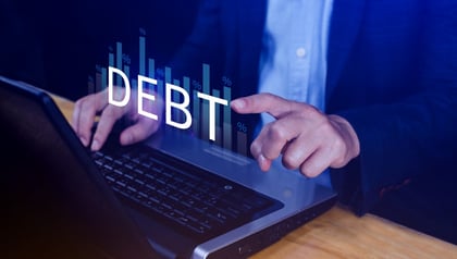 Businessman pointing at a glowing 'DEBT' graphic with rising percentage bar charts while typing on a laptop, representing technical debt and its growing cost