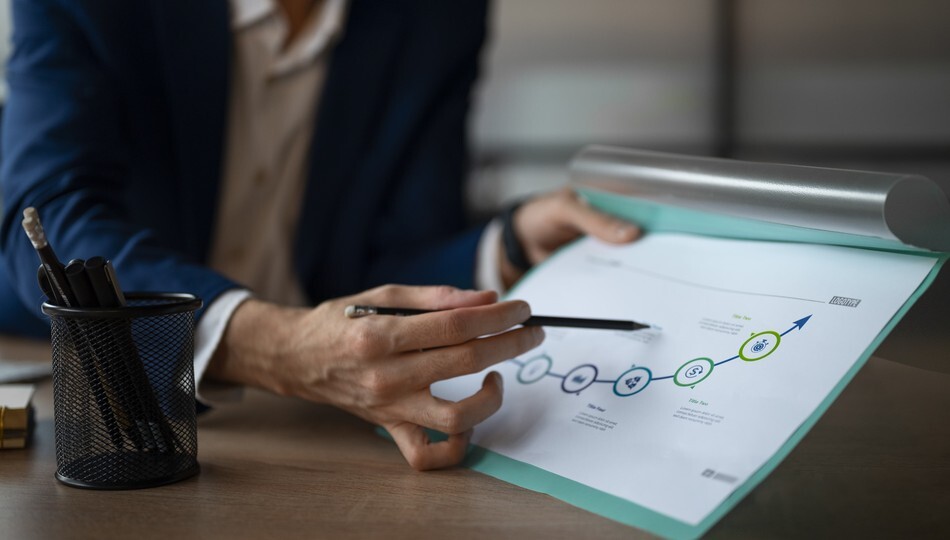 Businessman pointing pencil at a printed roadmap chart with milestone icons and an upward arrow.