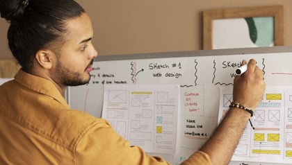 Man sketching wireframes on a whiteboard covered with web design sketches and sticky notes.