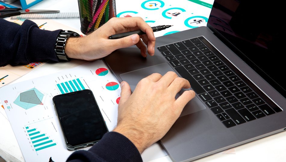 Hands typing on laptop beside printed charts, graphs, and a smartphone on a busy desk.