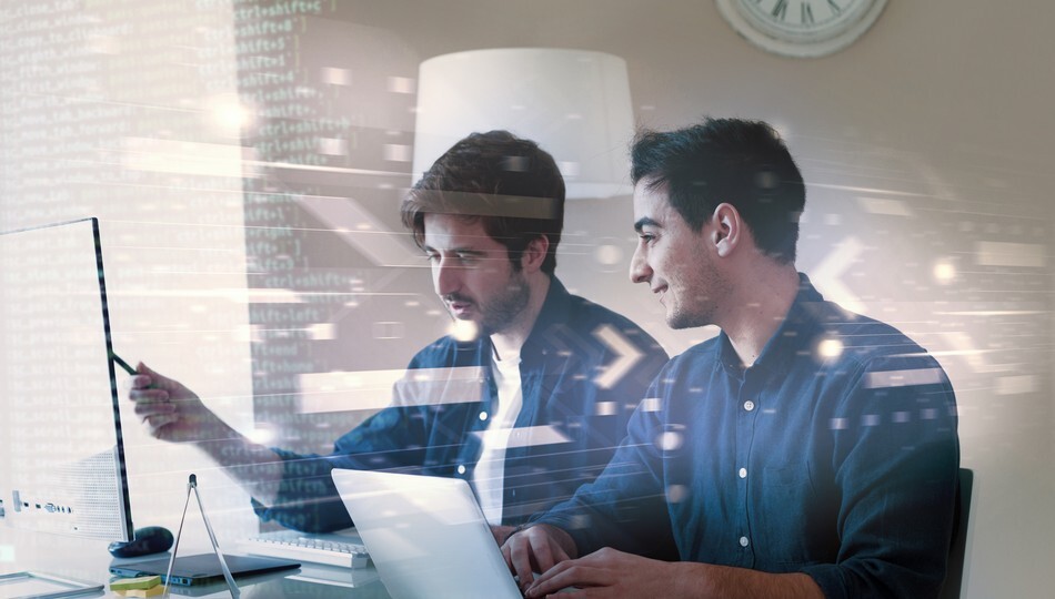 Two developers reviewing code on a laptop and monitor, representing enterprise software development collaboration.