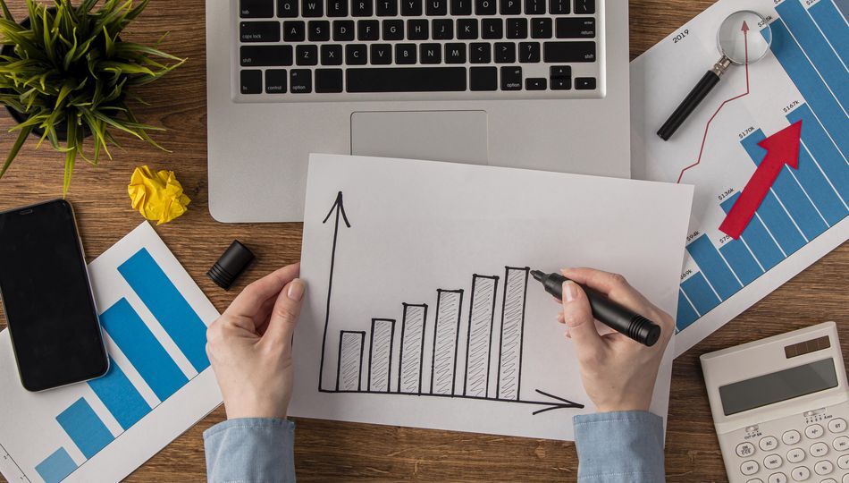 Hands sketching an upward growth chart on desk with financial charts, calculator, and laptop.