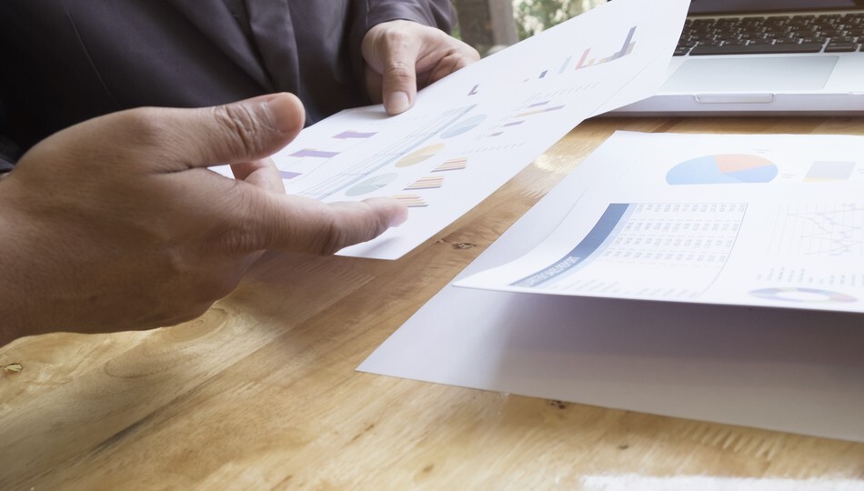 Hands reviewing printed charts and reports near a laptop, representing a product requirements document.