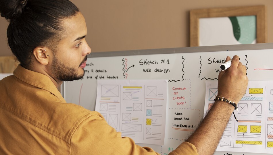 Man sketching wireframes on a whiteboard covered with web design sketches and sticky notes.